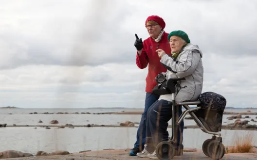 Ikäihminen istuu rollaattorin päällä meren rannalla laakealla kalliolla  ja katsoo yhdessä ulkoiluystävän kanssa maisemaa.