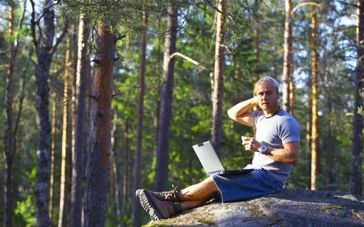 Mies istuu metsän keskellä kalliolla kannettava tietokone sylissä. 
