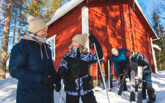 Nainen ja tyttö hymyilevät auringon paisteessa hankien keskellä. Mies ja poika huoltavat liukulumikenkiä taustalla.