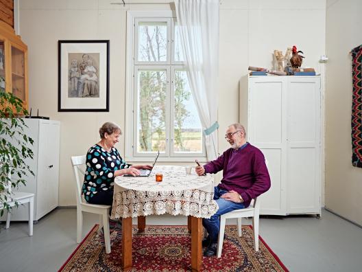 Ikääntyneet mies ja nainen istuvat pöydän ääressä. Nainen katsoo tietokonetta ja mies puhelinta.