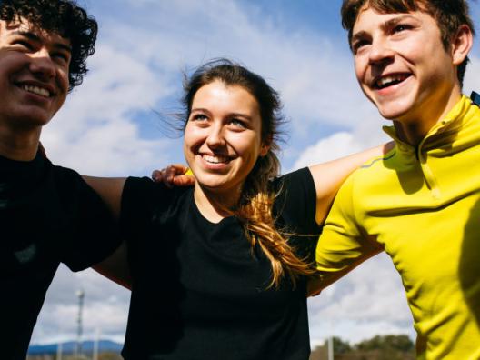 Kolme hymyilevää nuorta seisoo ulkona kädet toistensa hartioilla, aurinko paistaa ja taustalla näkyy sinistä taivasta.