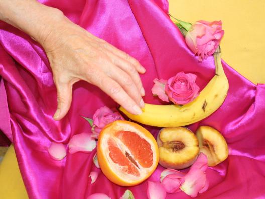Kuvassa on taustana pinkkiä ja keltaista kangas johon on aseteltu banaani, veriappelsiini ja pesikoita, joita kohden kurkottaa ikääntyneen naisen käsi