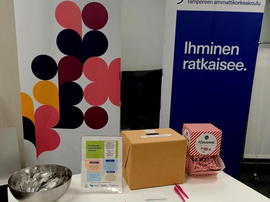 Esittelypöytä, jossa kaksi roll-up -mainosta: vasemmalla Pirkanmaan hyvinvointialueen värikäs graafinen banneri ja oikealla Tampereen ammattikorkeakoulun turkoosi banneri tekstillä Ihminen ratkaisee. Pöydällä on metallikulho, esitteitä, pinkkejä kyniä, pahvilaatikko ja karkkipussi.