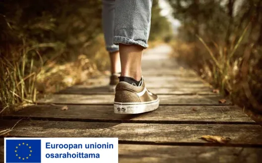 Askeleet puisella polulla, henkilön jalat näkyvät puisella alustalla. Kuvassa EU-lippulogo jossa teksti Euroopan unionin osarahoittama. 