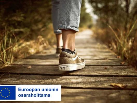 Askeleet puisella polulla, henkilön jalat näkyvät puisella alustalla. Kuvassa EU-lippulogo jossa teksti Euroopan unionin osarahoittama. 