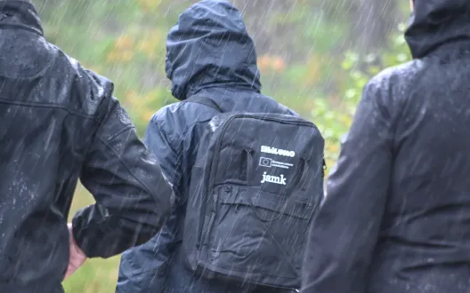 Kolme ihmistä ulkoilemassa syksyisessä sadesäässä, keskimmäisen ihmisen selässä on kirjastosta lainattava luontoreppu.