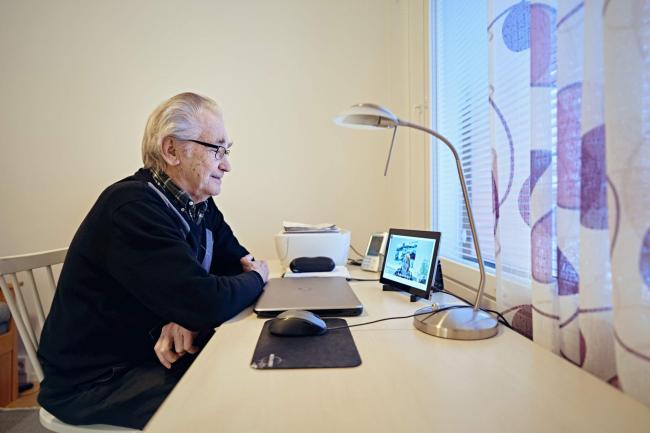 ikäihminen istuu pöydän ääressä ja katsoo kannettavan tietokoneen näyttöä. 
