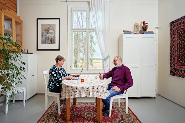 Ikääntyneet mies ja nainen istuvat pöydän ääressä. Nainen katsoo tietokonetta ja mies puhelinta.