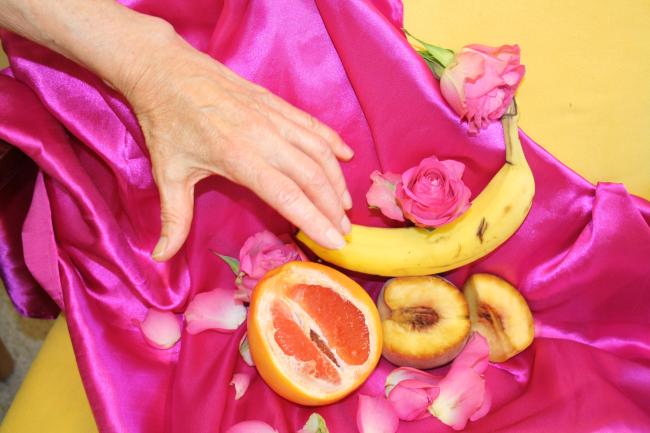 Kuvassa on taustana pinkkiä ja keltaista kangas johon on aseteltu banaani, veriappelsiini ja pesikoita, joita kohden kurkottaa ikääntyneen naisen käsi
