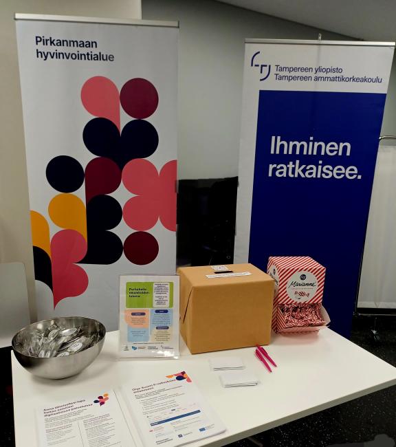Esittelypöytä, jossa kaksi roll-up -mainosta: vasemmalla Pirkanmaan hyvinvointialueen värikäs graafinen banneri ja oikealla Tampereen ammattikorkeakoulun turkoosi banneri tekstillä Ihminen ratkaisee. Pöydällä on metallikulho, esitteitä, pinkkejä kyniä, pahvilaatikko ja karkkipussi.