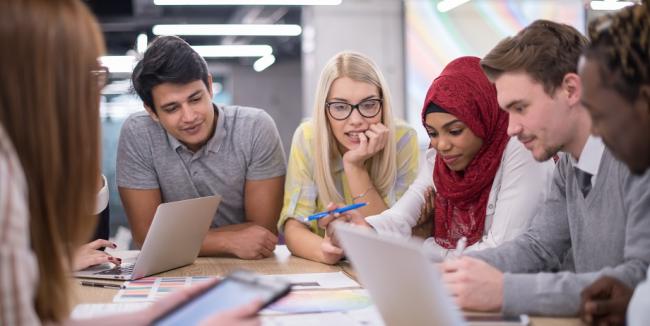 Kehittäjät seisovat pöydän ääressä keskustellen ja pohtien. Pöydällä on papereita ja tietokone.